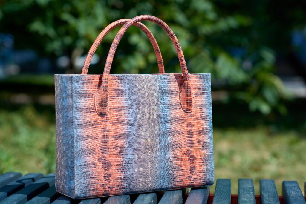 close up fashionable woman s bag with snake skin imitation stands blue park bench bag was made blue pink grey colors also it has comfortable handles 7502 5648